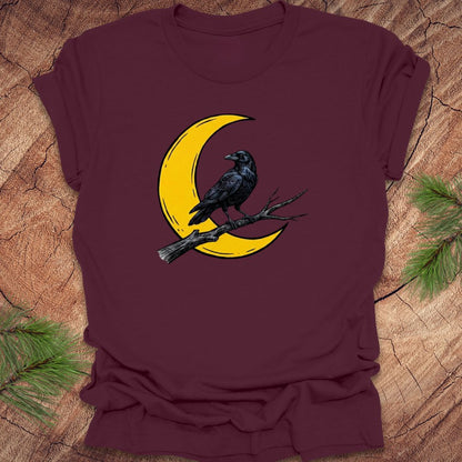 Maroon t-shirt with a yellow crescent moon and black crow design on a wooden surface.