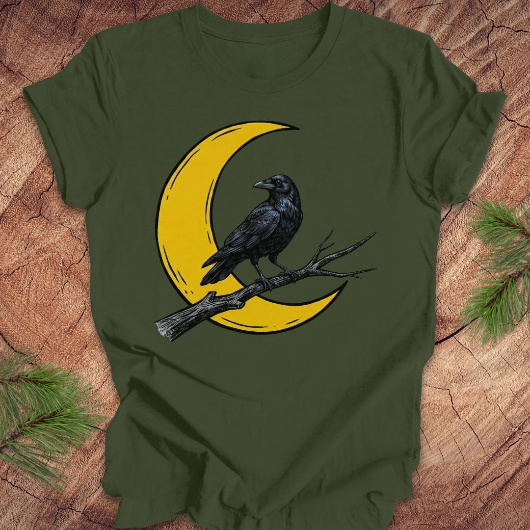 An olive green short-sleeve t-shirt with a graphic of a black raven perched on a branch in front of a yellow crescent moon, laid flat on a rustic wood background with pine needles in the corners.