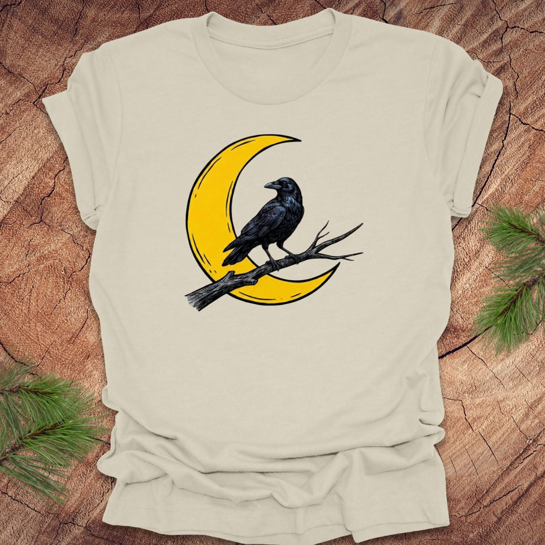 Beige t-shirt with a yellow crescent moon and black crow design on a wooden surface.