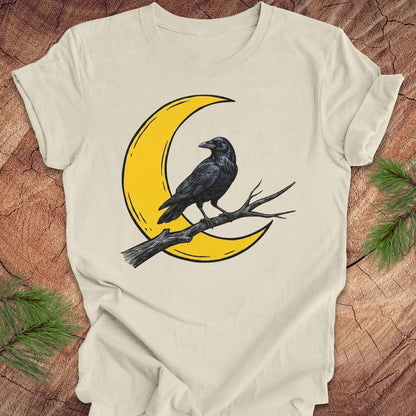 A cream-colored short-sleeve t-shirt with a graphic of a black raven perched on a yellow crescent moon on a branch, laid flat on a rustic wood background with pine needles in the corners.