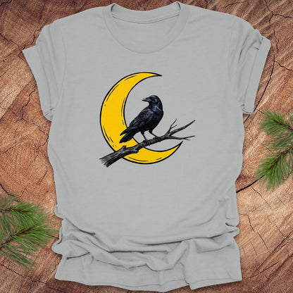 Gray t-shirt with a yellow crescent moon and black crow design on a wooden surface.