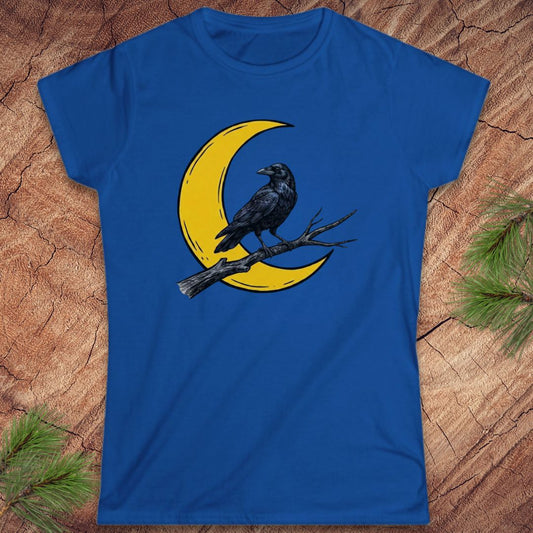A royal blue short-sleeve t-shirt with a graphic of a black raven perched on a branch in front of a yellow crescent moon, laid flat on a textured wood background with pine needles in the corners.