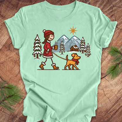 Light green t-shirt with a graphic of a person walking a dog in a winter scene on a wooden surface.