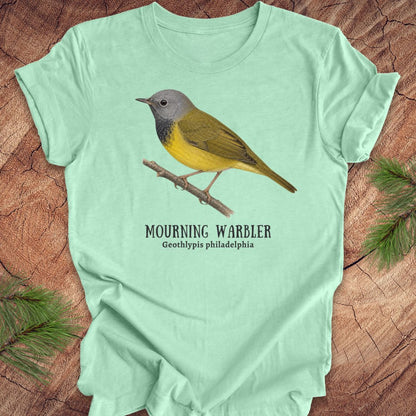 Light green t-shirt with a mourning warbler illustration and text on a wooden surface