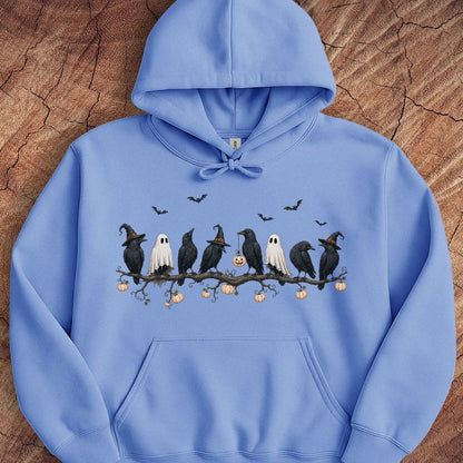 A light blue pullover hoodie featuring a Halloween graphic of black crows, white ghosts, and pumpkins on a branch, laid flat on a textured wood background.