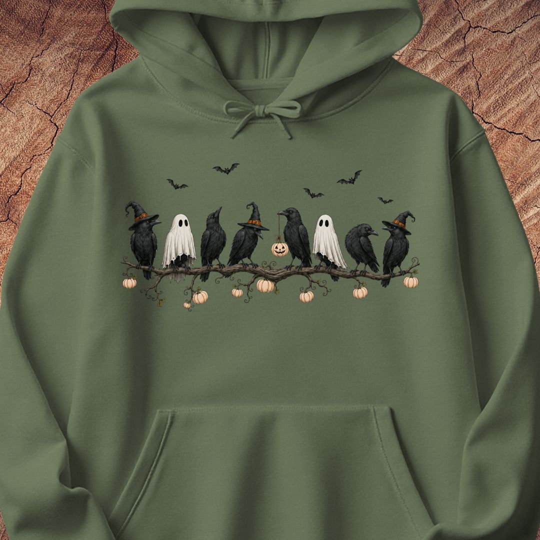 An olive green pullover hoodie featuring a Halloween graphic of crows, ghosts, pumpkins, and bats on a branch, displayed against a textured wood background.
