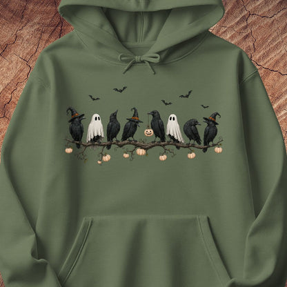An olive green pullover hoodie featuring a Halloween graphic of crows, ghosts, pumpkins, and bats on a branch, displayed against a textured wood background.