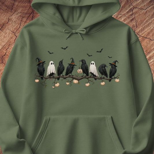 An olive green pullover hoodie featuring a Halloween graphic of crows, ghosts, pumpkins, and bats on a branch, displayed against a textured wood background.
