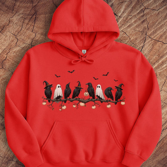 A red pullover hoodie with a Halloween-themed graphic of crows, ghosts, and bats on a branch, laid flat on a textured wood background.
