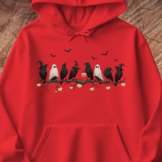 A bright red pullover hoodie featuring a Halloween graphic of crows, ghosts, and pumpkins on a branch, displayed against a textured wood background.