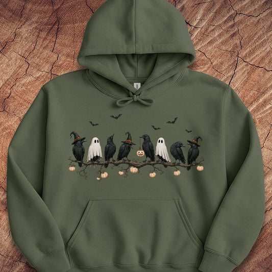 An olive green pullover hoodie featuring a Halloween-themed graphic print of crows, ghosts, pumpkins, and bats on a branch, displayed flat against a textured wood background.