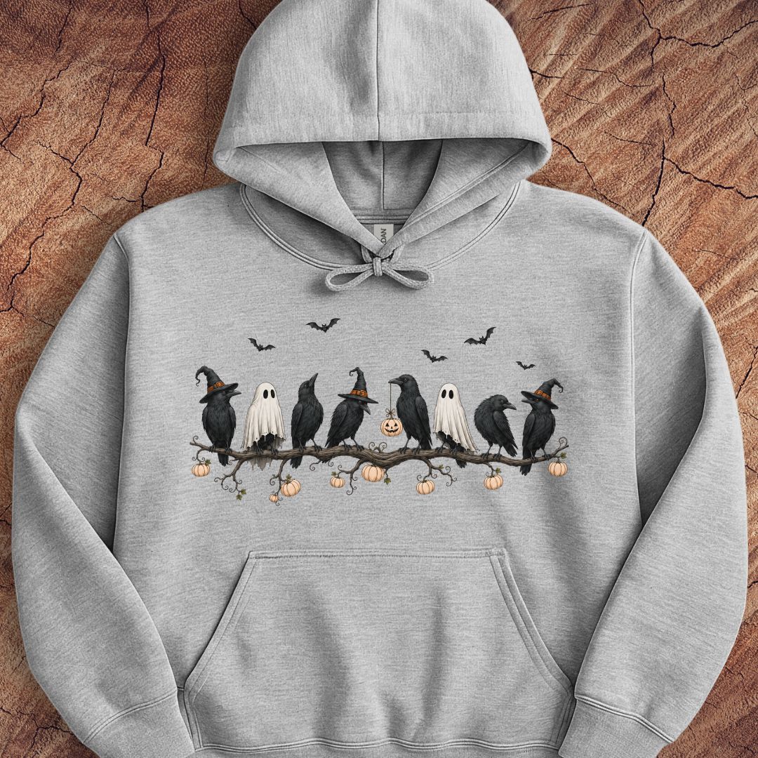 A light gray pullover hoodie featuring a Halloween-themed graphic print of crows, ghosts, and pumpkins on a branch, displayed flat against a textured wood background.