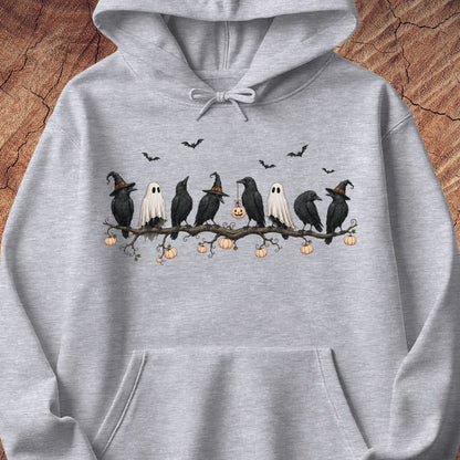 A light heather gray hoodie featuring a Halloween graphic print of crows, ghosts, and pumpkins on a branch, displayed against a rustic wood background.