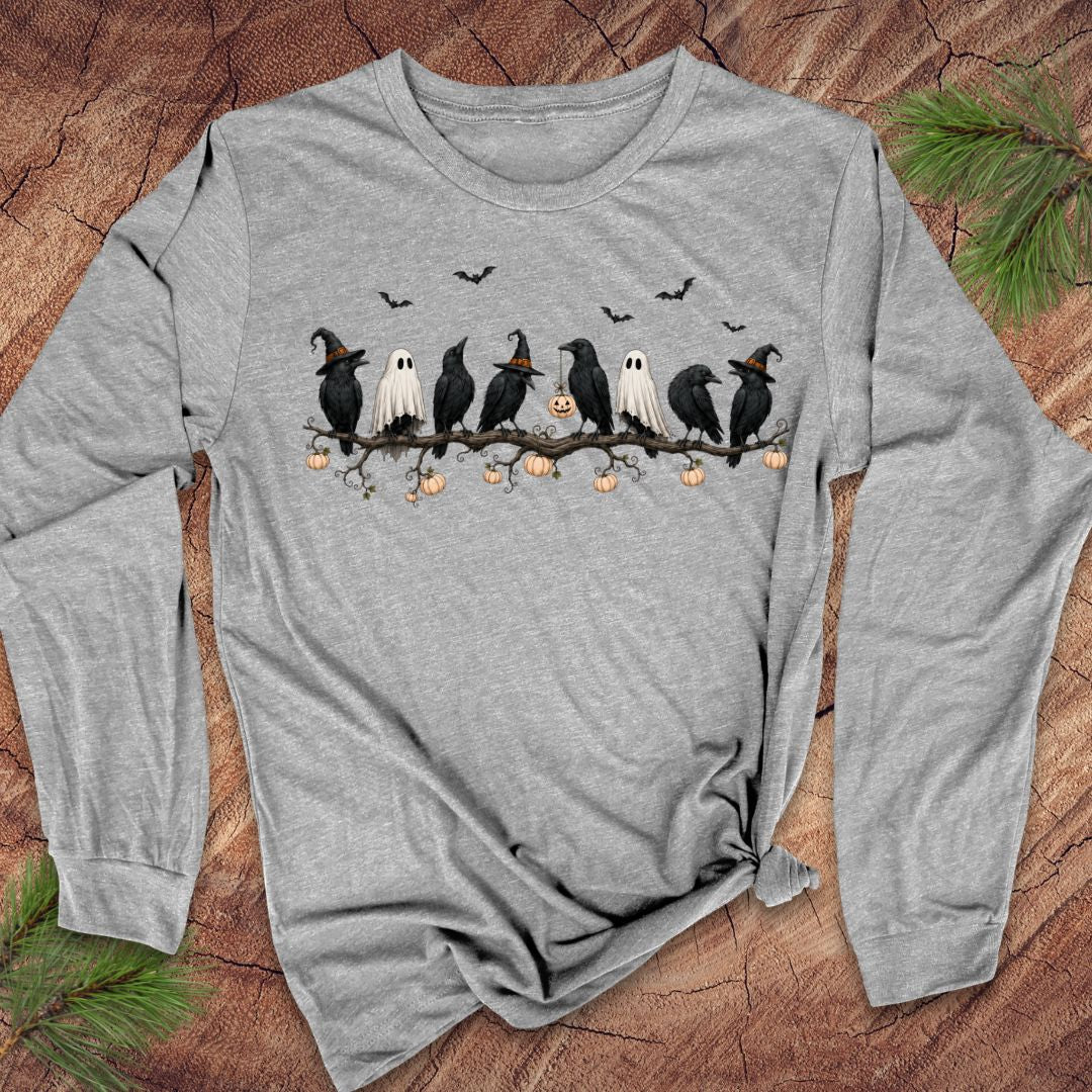 A heather gray long-sleeve t-shirt with a Halloween graphic of five crows, two ghosts, and pumpkins on a branch, laid flat on a rustic wood background with pine needles.