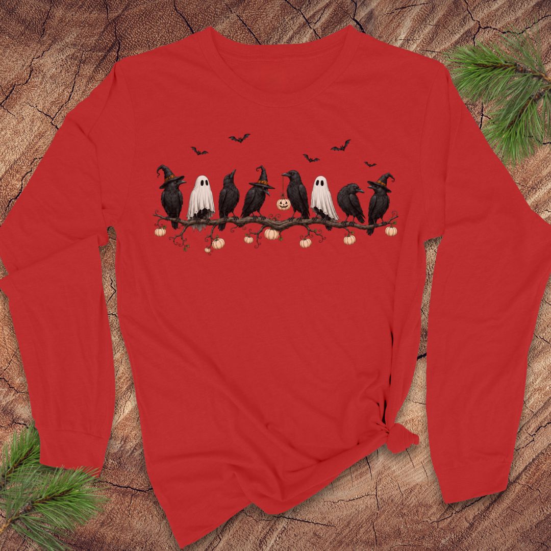 A red long-sleeve t-shirt with a Halloween graphic of crows, ghosts, and pumpkins, laid flat on a rustic wood background with pine branches.