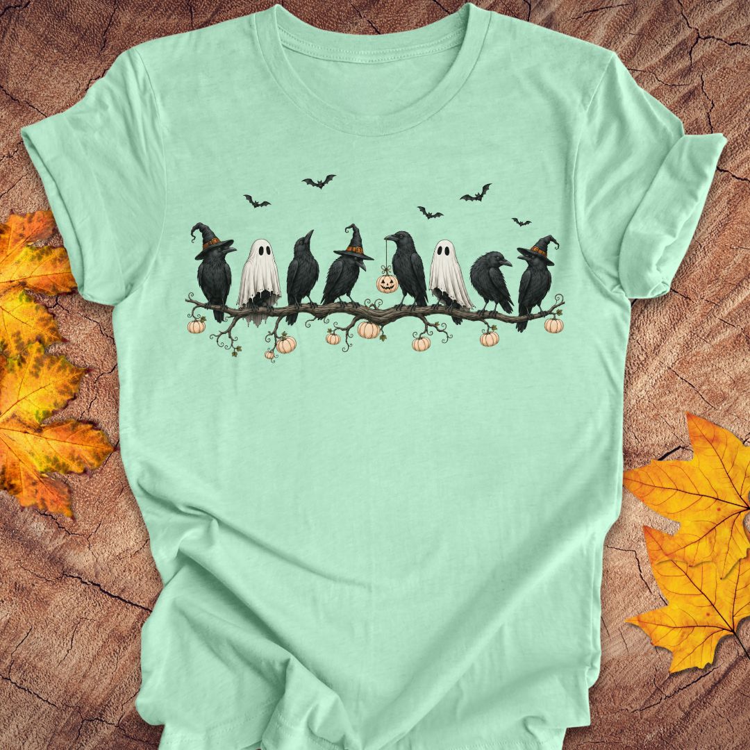 A mint green short-sleeve t-shirt with a Halloween graphic print of crows, ghosts, pumpkins, and bats, styled flat on a wood background with autumn leaves.