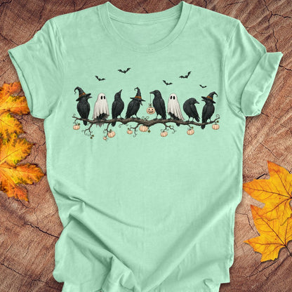 A mint green short-sleeve t-shirt with a Halloween graphic print of crows, ghosts, pumpkins, and bats, styled flat on a wood background with autumn leaves.