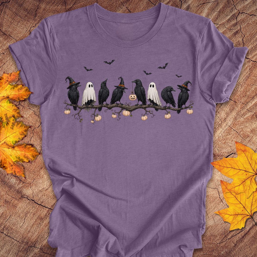 A purple short-sleeve crewneck t-shirt featuring a Halloween graphic of crows, ghosts, pumpkins, and bats on a branch, laid flat on a rustic wood background with autumn leaves.