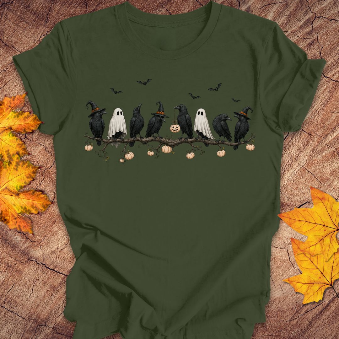 An olive green short-sleeve t-shirt with a Halloween graphic print, laid flat on a rustic wood background with autumn leaves. The graphic shows black crows and ghosts on a branch with pumpkins and bats.