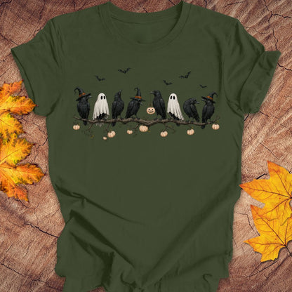 An olive green short-sleeve t-shirt with a Halloween graphic print, laid flat on a rustic wood background with autumn leaves. The graphic shows black crows and ghosts on a branch with pumpkins and bats.