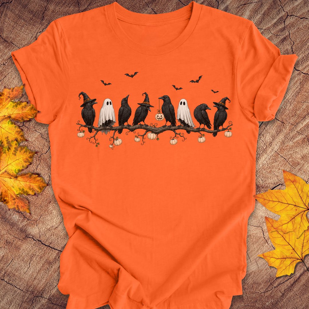 An orange short-sleeve crewneck t-shirt featuring a graphic of crows wearing witch hats and white ghosts on a branch with pumpkins, laid flat on a rustic wood background with autumn leaves.