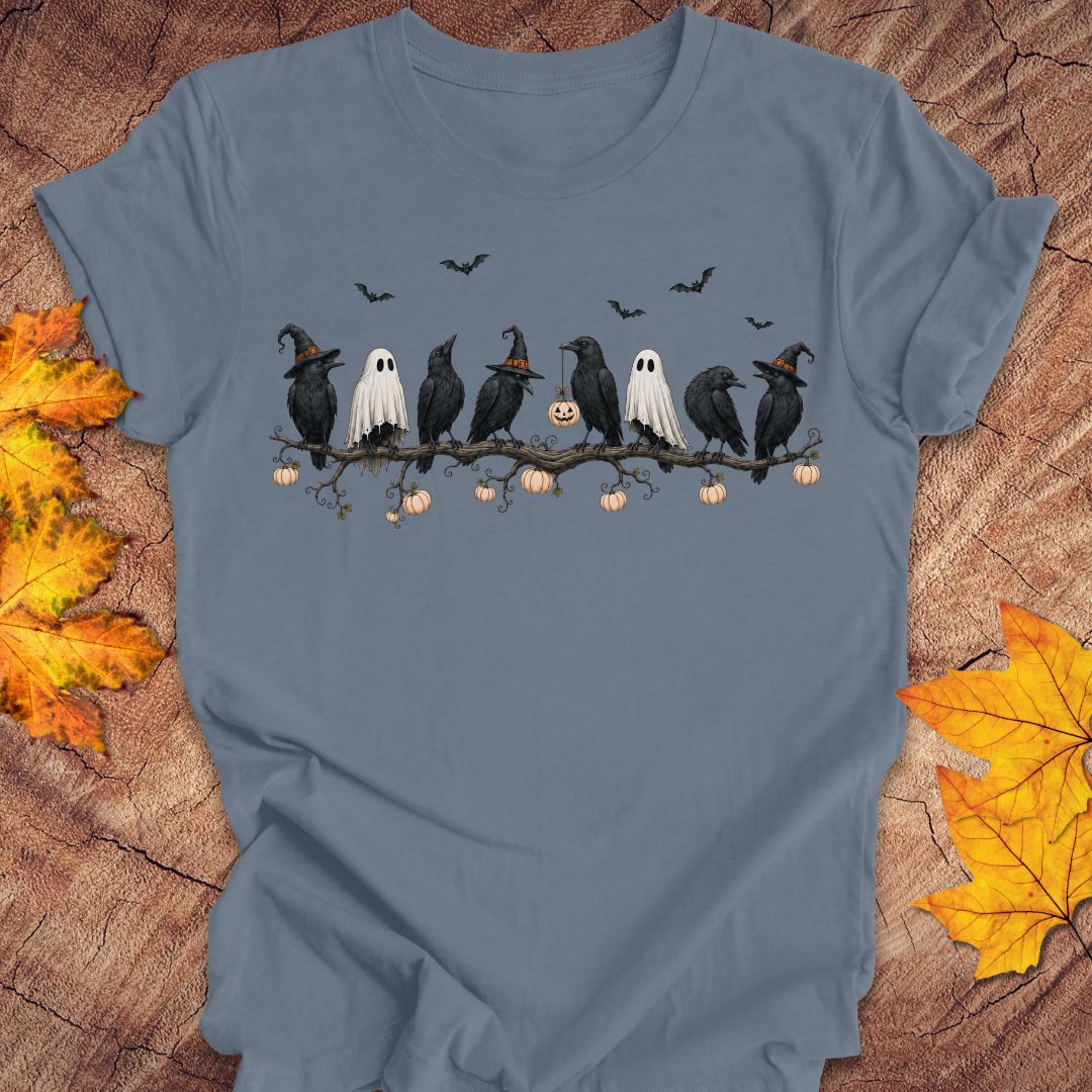A slate blue short-sleeve t-shirt with a Halloween graphic of black crows, white ghosts, and pumpkins on a branch, displayed flat on a rustic wood background with autumn leaves.