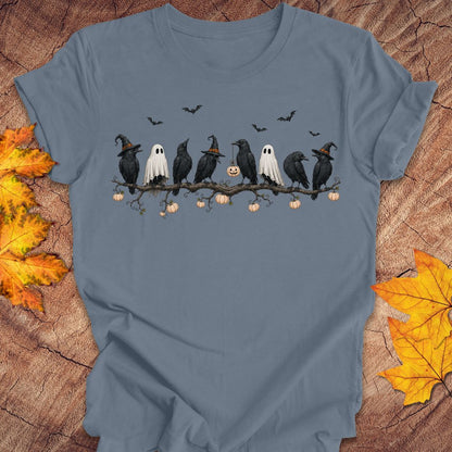 A slate blue short-sleeve t-shirt with a Halloween graphic of black crows, white ghosts, and pumpkins on a branch, displayed flat on a rustic wood background with autumn leaves.