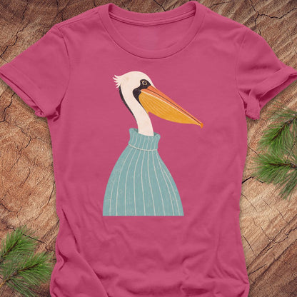 A pink short-sleeve t-shirt laid flat on a rustic wood surface, featuring a stylized illustration of a white pelican with a yellow-orange beak and a light blue striped sweater.