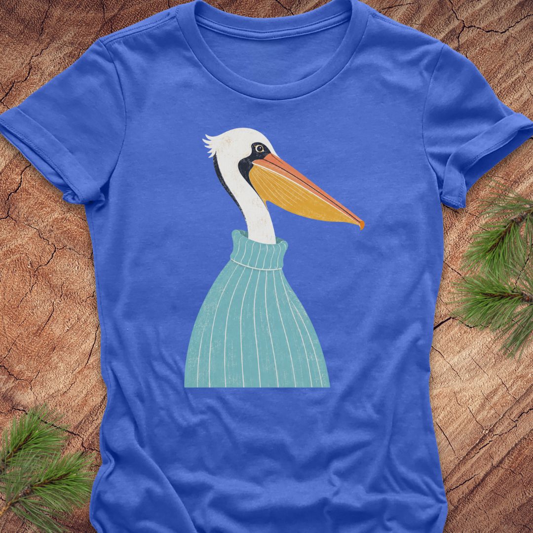 A royal blue short-sleeve t-shirt laid flat on a textured wood surface, featuring a stylized graphic of a white pelican with a yellow-orange beak and a light green striped sweater. Pine needles are visible in the corners as flat-lay props.
