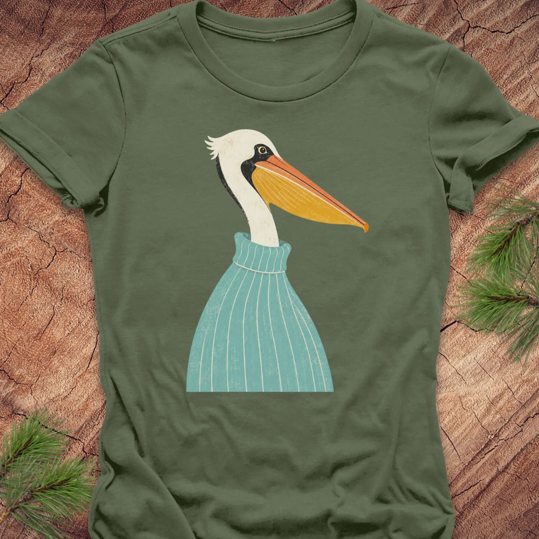 An olive green short-sleeve t-shirt featuring a stylized graphic of a white pelican wearing a light blue striped sweater, laid flat on a rustic wood background with pine needles.