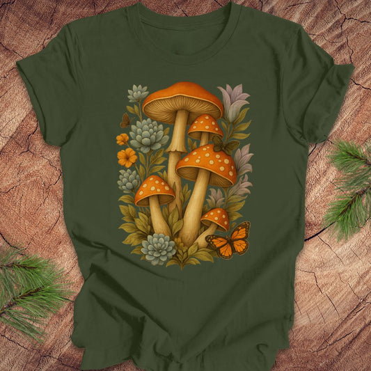 An olive green short-sleeve t-shirt with a colorful graphic of orange mushrooms, green succulents, yellow flowers, and an orange butterfly, laid flat on a textured wood background with pine branches in the corners.