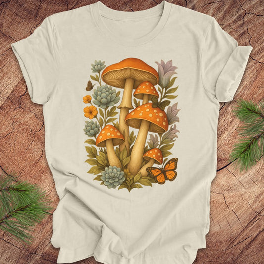 An beige short-sleeve t-shirt with a detailed graphic of orange mushrooms, green succulents, and an orange butterfly, laid flat on a rustic wood background with pine branches.