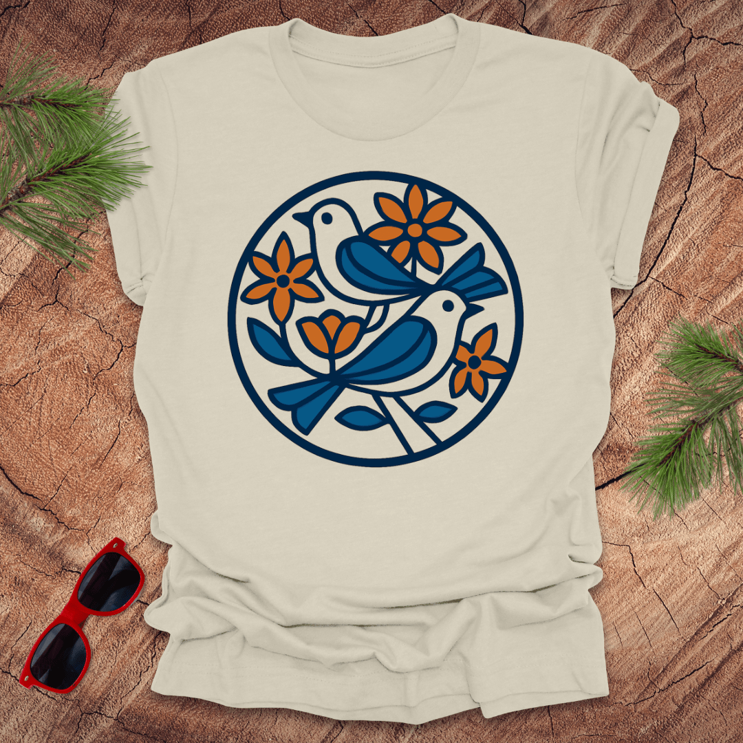Beige t-shirt with a blue and orange floral and bird design on a wooden surface with sunglasses and greenery.
