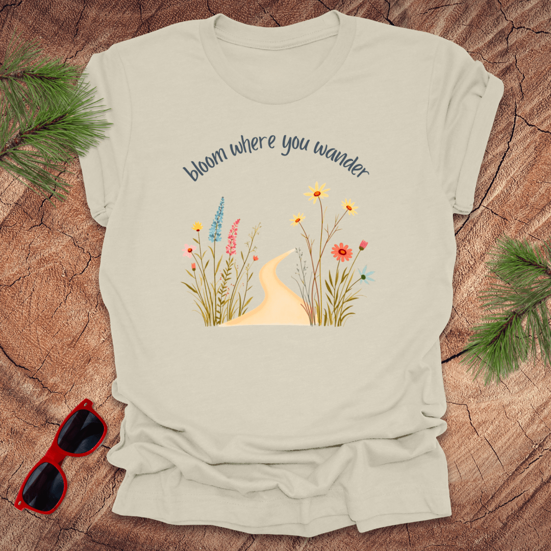 beige t-shirt with a graphic that includes a floral design and the text 'bloom where you wander', accompanied by  flowers on a wooden background with pine needles and red sunglasses to the side.