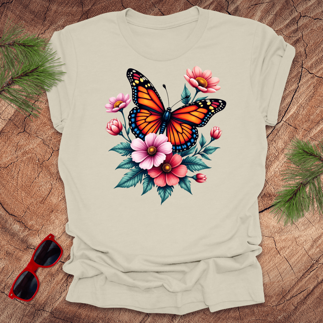 A beige t-shirt with a vibrant print of a butterfly surrounded by pink and red flowers, displayed on a wooden surface with green leaves and red sunglasses to the side.