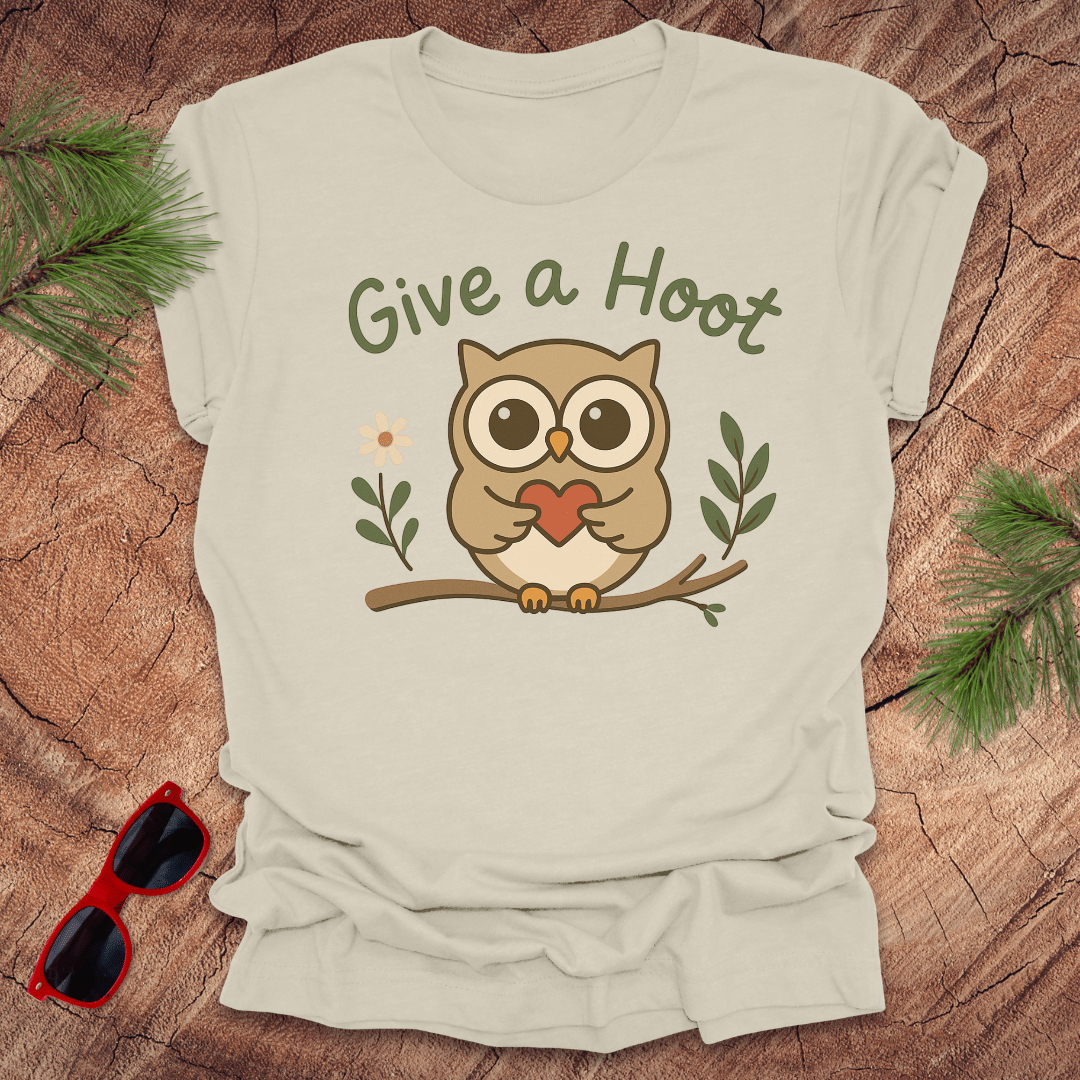 Beige t-shirt with owl graphic and 'Give a Hoot' text on a wooden surface with sunglasses and greenery.