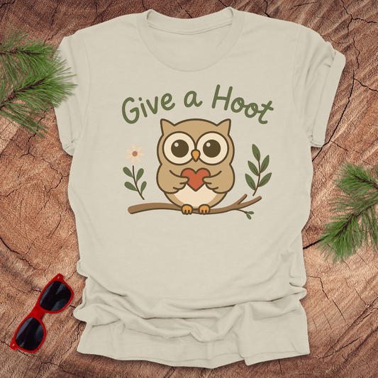 Beige t-shirt with owl graphic and 'Give a Hoot' text on a wooden surface with sunglasses and greenery.