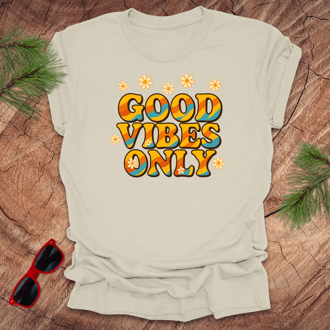 Beige t-shirt with 'Good Vibes Only' text on a wooden surface with sunglasses and greenery.