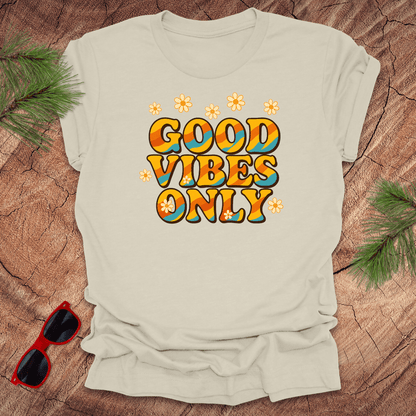 Beige t-shirt with 'Good Vibes Only' text on a wooden surface with sunglasses and greenery.
