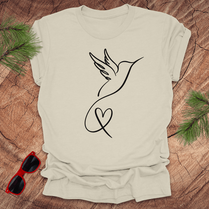 Beige t-shirt with hummingbird and heart design on a wooden surface with sunglasses and greenery.