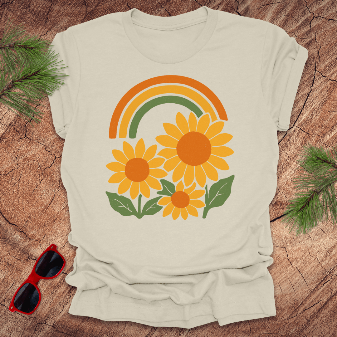 Beige t-shirt with a rainbow and sunflower design on a wooden surface with sunglasses and greenery.