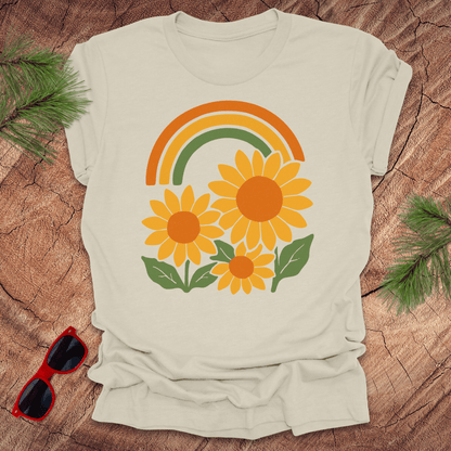 Beige t-shirt with a rainbow and sunflower design on a wooden surface with sunglasses and greenery.