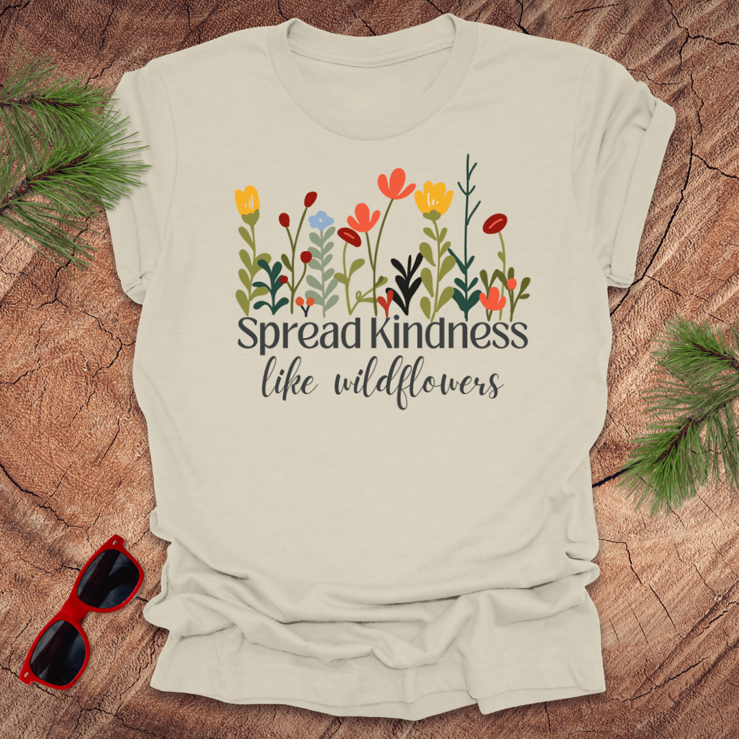 Beige t-shirt with floral design and 'Spread kindness like wildflowers' text on a wooden surface with sunglasses and greenery.