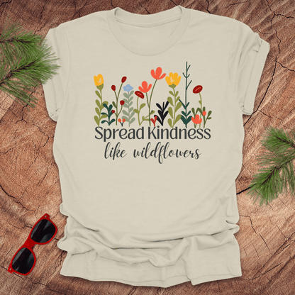 Beige t-shirt with floral design and 'Spread kindness like wildflowers' text on a wooden surface with sunglasses and greenery.