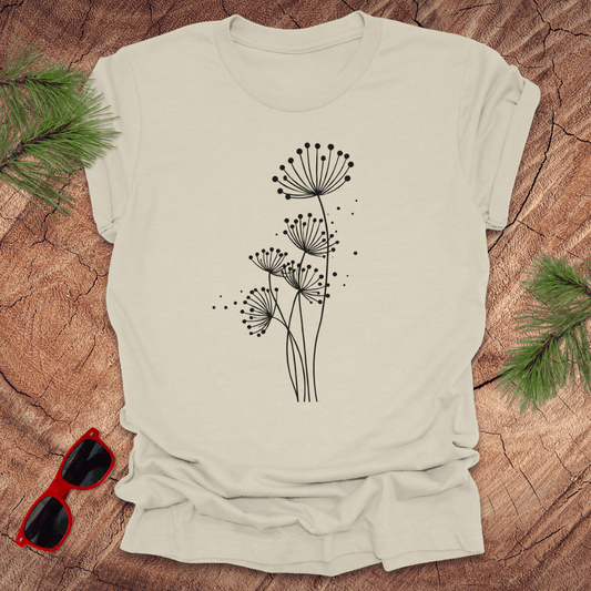 Beige t-shirt with black floral dandelion design on a wooden surface with sunglasses and greenery.