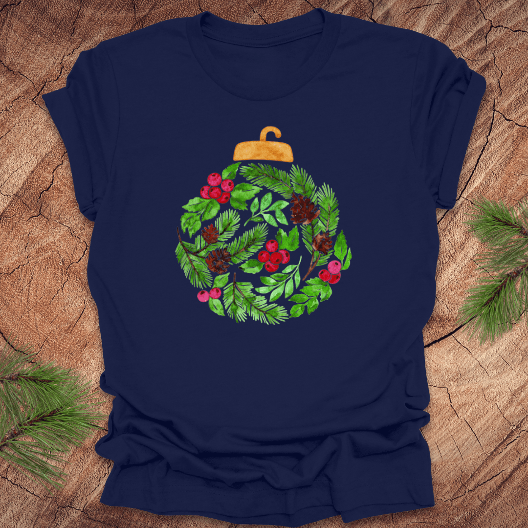 Navy blue t-shirt with a Christmas ornament design featuring green leaves, red berries, and gold accents on a wooden surface.