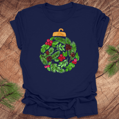 Navy blue t-shirt with a Christmas ornament design featuring green leaves, red berries, and gold accents on a wooden surface.