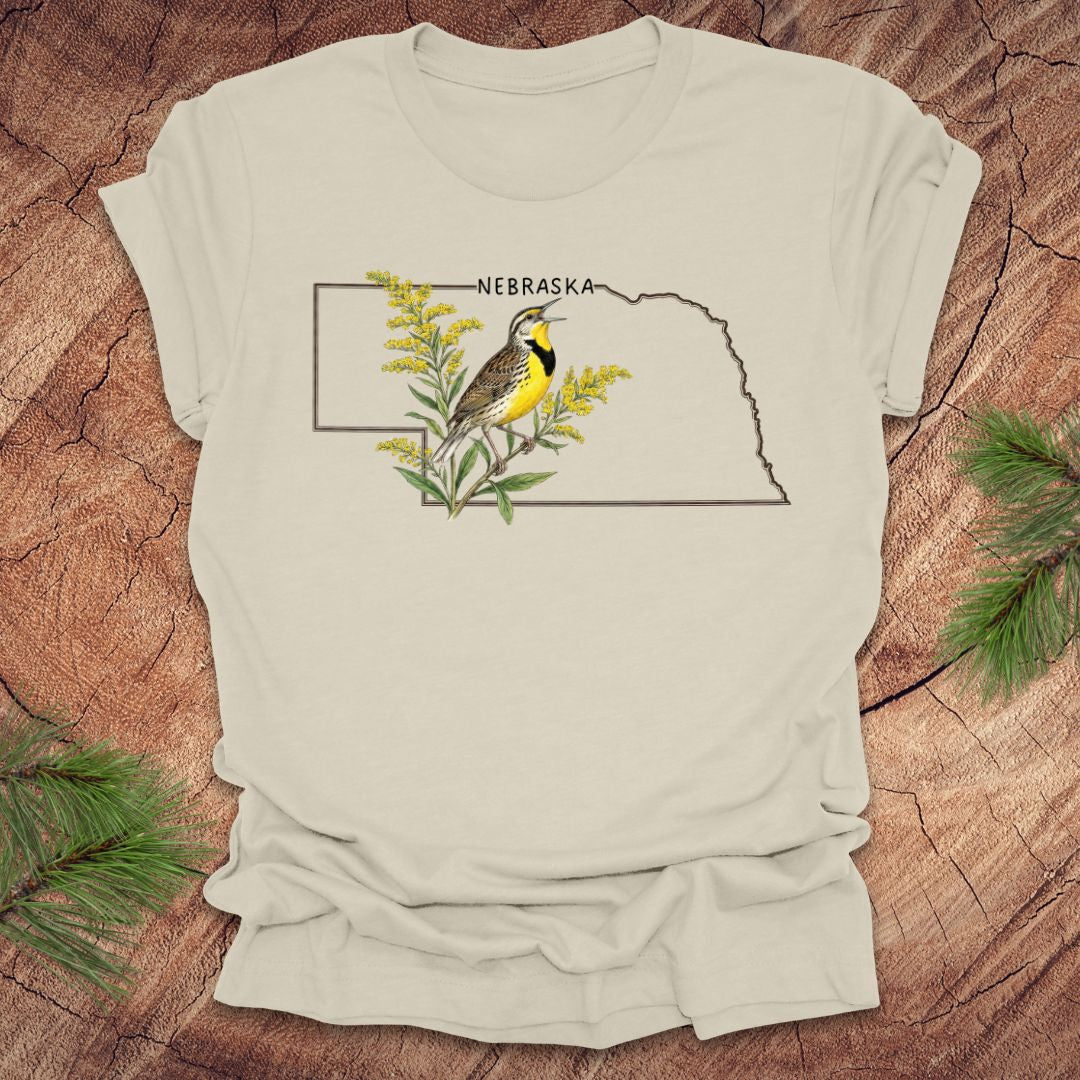 Beige t-shirt with the outline of the state of Nebraska and a  graphic of a meadowlark and goldenrod inside on a wooden surface