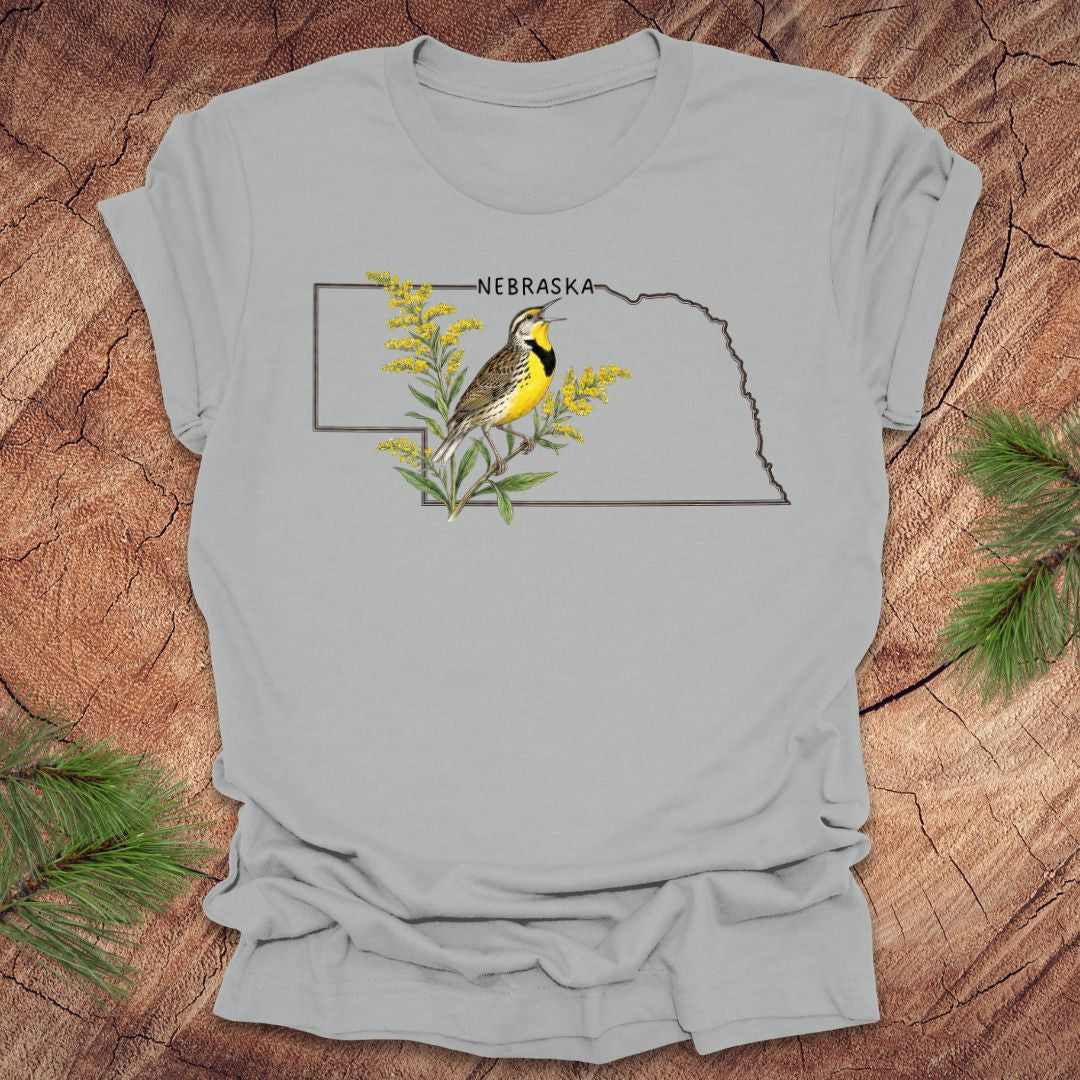 Silver t-shirt with the outline of the state of Nebraska and a  graphic of a meadowlark and goldenrod inside on a wooden surface