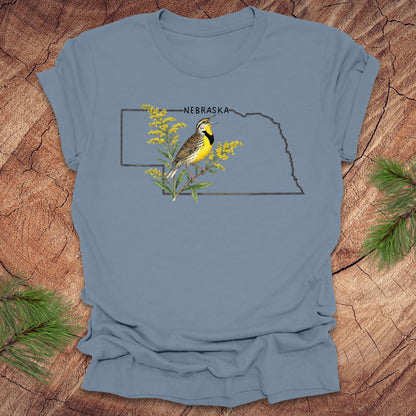 Steel blue t-shirt with the outline of the state of Nebraska and a  graphic of a meadowlark and goldenrod inside on a wooden surface